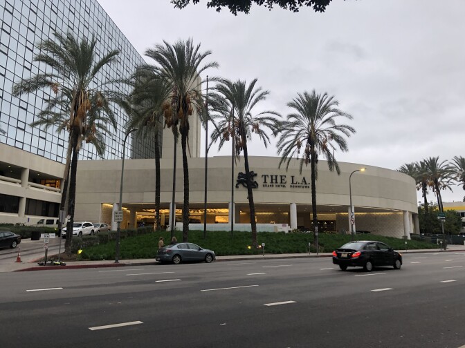 The facade of a building is rounded with the words The L.A. visible above a covered drive. Palm trees are in front and behind and to the left is a multi-story building with a mirrored glass front