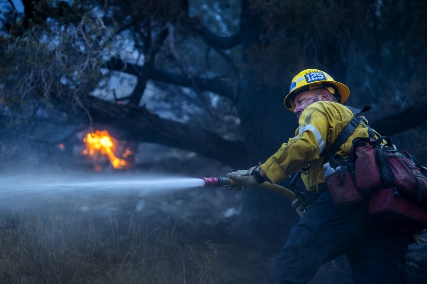 A firefighter wearing a yellow hard hat and yellow jacket is directing a hose of water to his front left, out of view of the camera. Red and orange flames can be seen burning in the background amid trees. The sky is sort of dark and hazy.