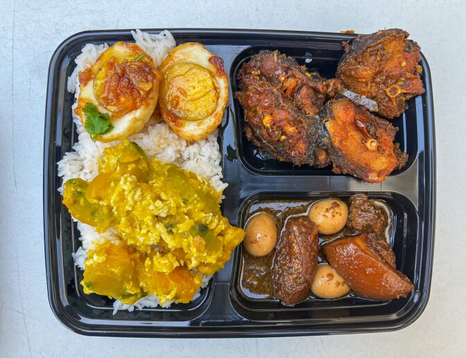 A black plastic to-go tray with three different compartments, each filled with varying types of food. On the left is a bed of cooked white rice containing chunks of an orange squash-like vegetable in a yellow sauce towards the bottom of the tray. On top of the rice are two hard-boiled eggs with a light reddish-brown sauce. The right side has two smaller sections. The top section is filled with large chunks of sliced fish in a dark red sauce, while the bottom section contains large chunks of brown cooked meat and small quail eggs in a light brown liquid sauce.