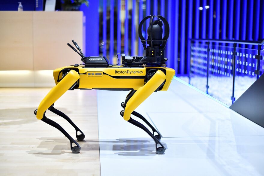 A four-legged robot has yellow legs and is shaped like a dog. A camera sits where a head would be.