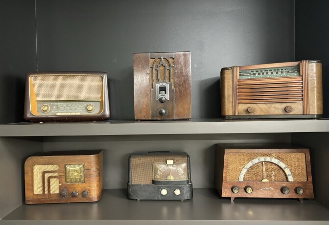 Six vintage radios sit on two shelves: There are three on the top shelf and three on the shelf below it, all of varying models.