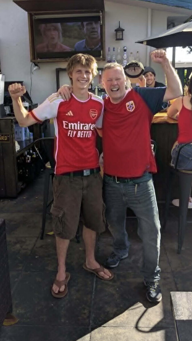 A teenage boy and a man with light skin tone both wear red and stand with their arms around one-another and a fist in the air. 