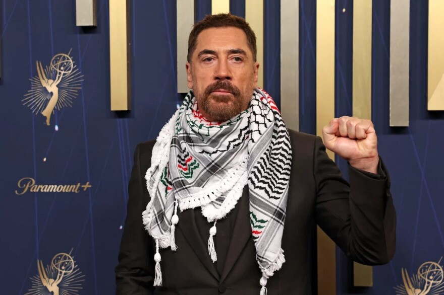 a man in a suit and a scarf stands against a blue and gold background and holds up a single fist