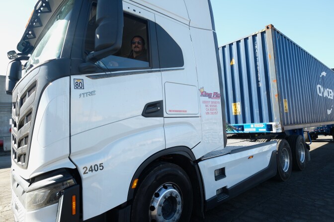 A large white flat-faced big rig truck with a blue shipping container on its trailer under a bright blue sky. 