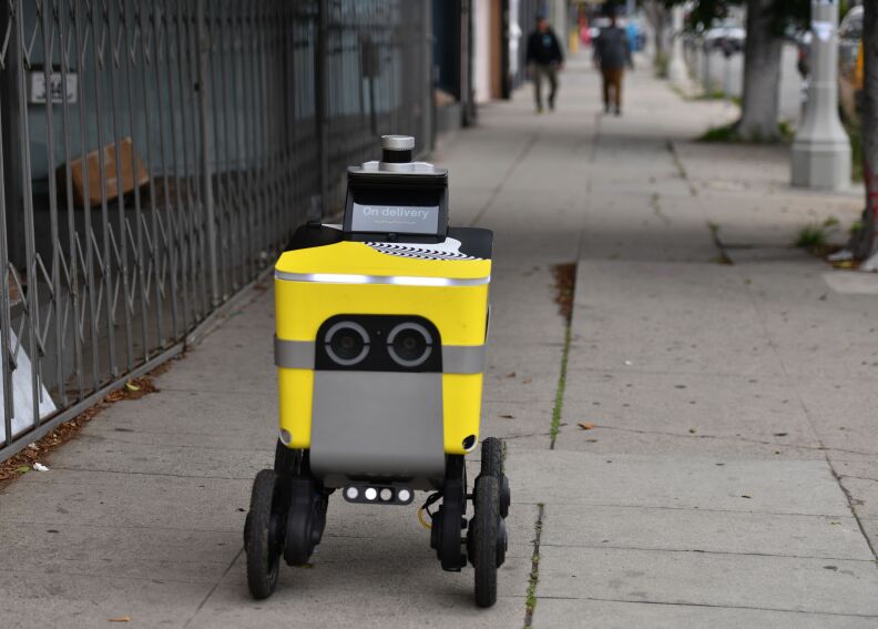A four-wheeled delivery robot rolls on a sidewalk in Los Angeles as people approach in the distance.
