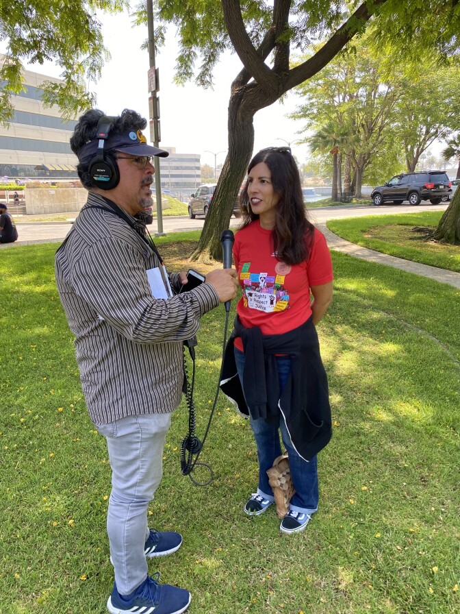 Two people talk in a park, under trees.