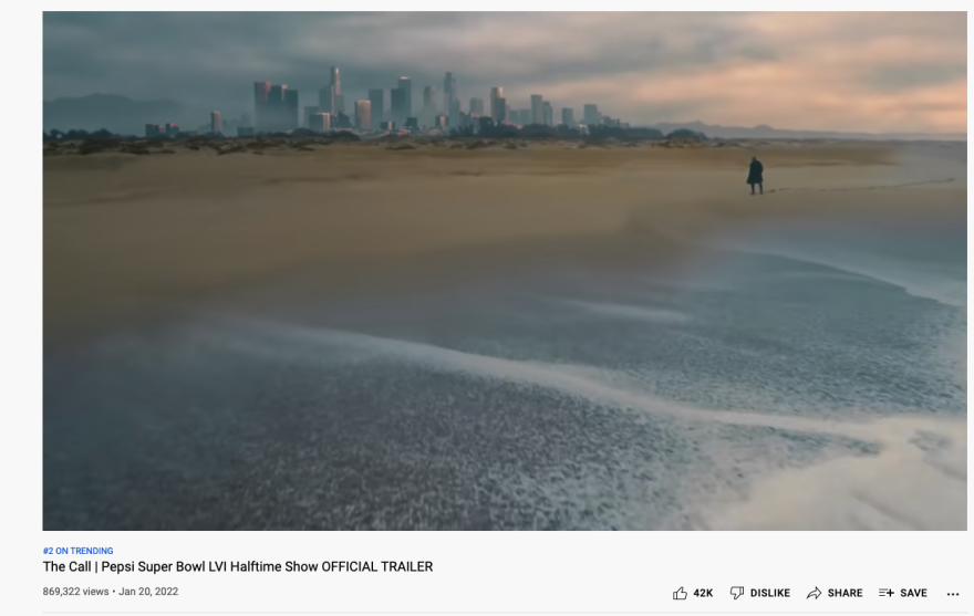A man in a dark coat walks along the beach with the skyline of downtown Los Angeles looming nearby.