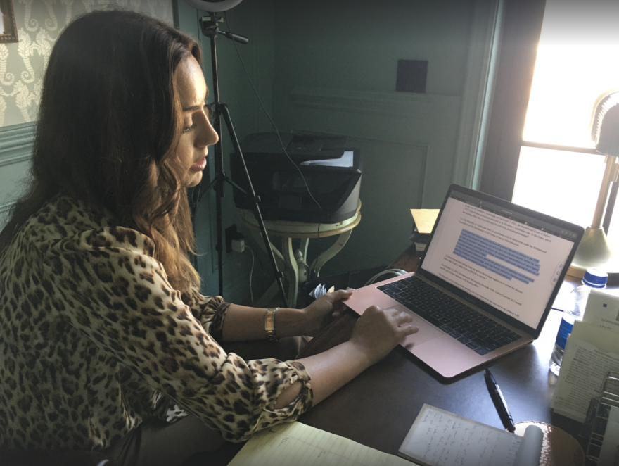 A woman sits at a laptop in a room lit by a bright window to one side.