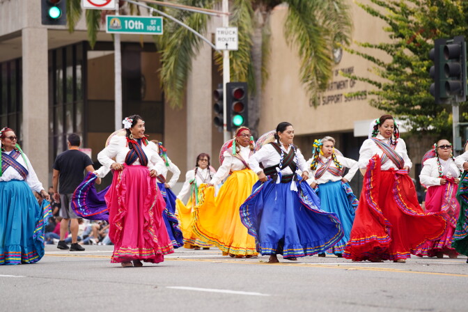 Folkorico dancers wearing skirts in varying colors perform in the street during a parade.