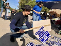 Man crouches to pick up strike signs that say, UAW, on strike, unfair labor practice.