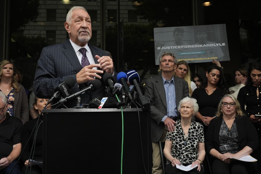 An older man in a suit and tie speaks into a microphone while a crowd of people behind him observes.
