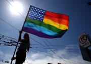 A person, who's blacked out in silhouette against the bright sun, holds a large Pride flag, which is the American flag filled in with rainbow colors insted of red and white stripes.