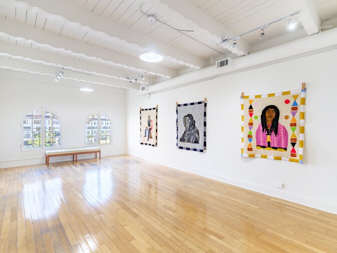 A bright white gallery space with shiny wood floors, three colorful square quilts on one wall, each of one person. On another wall are 2 windows and a bench.