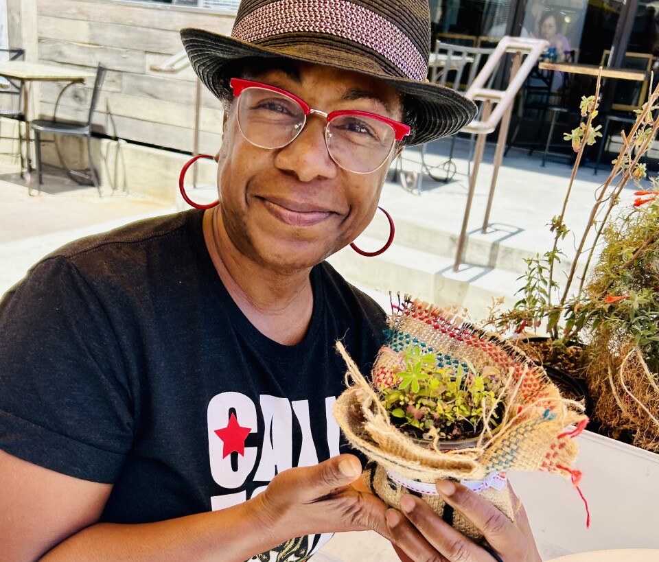 A woman wears a hat, red-framed glasses and a dark T-shirt while holding a plant in a pot.