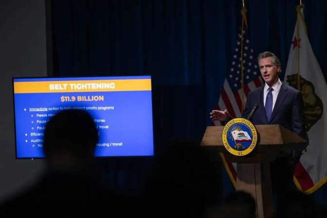 A light-skinned man with gray hair wearing a suit stands at a podium talking to a crowd. A television screen with a powerpoint is to his side. A shadow of a person covers part of the screen.