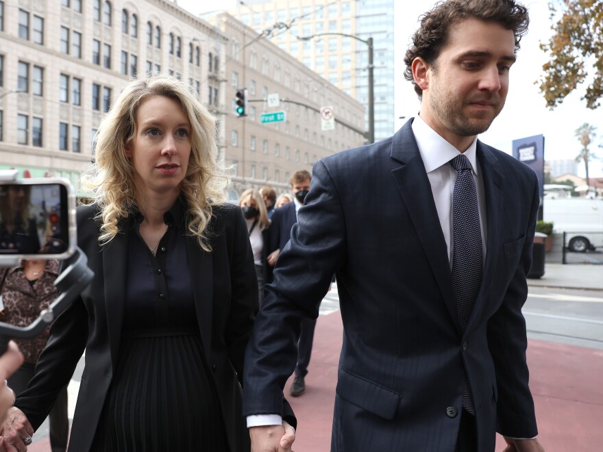 A white woman with blond hair wears dark clothing and holds the hand of a white man in a dark suit.