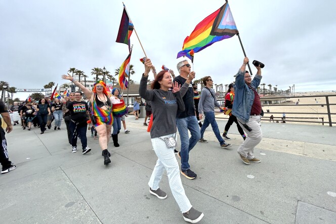 A crowd of peope march on the pier with their hands in the air with pride flags. Many people have rainbow colors on their clothing in some way.