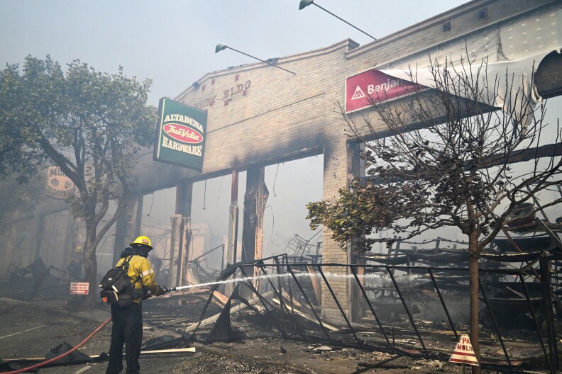 A firefighter hoses down the frame of a brick structure that has mostly burned down. There's a green sign hanging at the entrance that reads "Altadena Hardware."
