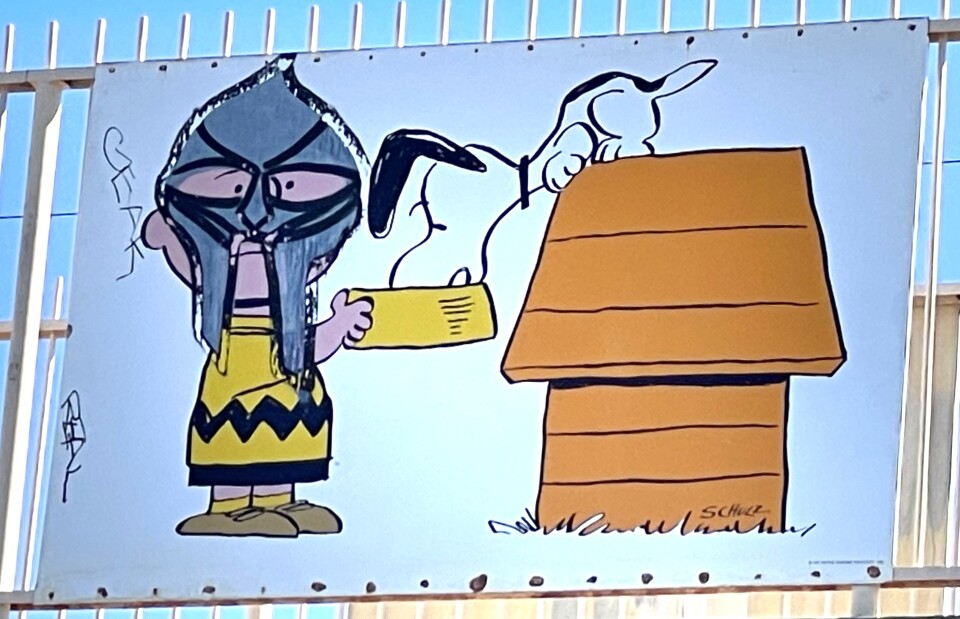 A white panel, featuring characters from the Peanuts comics, is affixed to the tan railings of a bridge. The panel depicts a cartoon boy wearing a yellow and black striped shirt and black shorts, with a gray mask drawn over its face after the fact. The boy is raising his arms to lift a yellow bowl towards his black and white dog that is sitting on top of a brown dog house. 