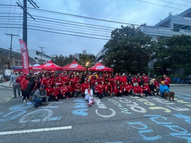 About 50 people stand before a set of tents, posing for a photograph. Most are wearing red, long-sleeved tops. The street before them has names like "Olga," "Ana," and "Jose" written in chalk. 