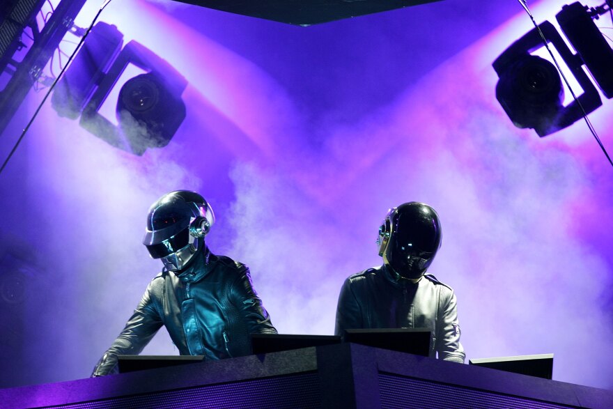 The band Daft Punk, wearing robotic helmets, stand at DJ consoles under purple lights.