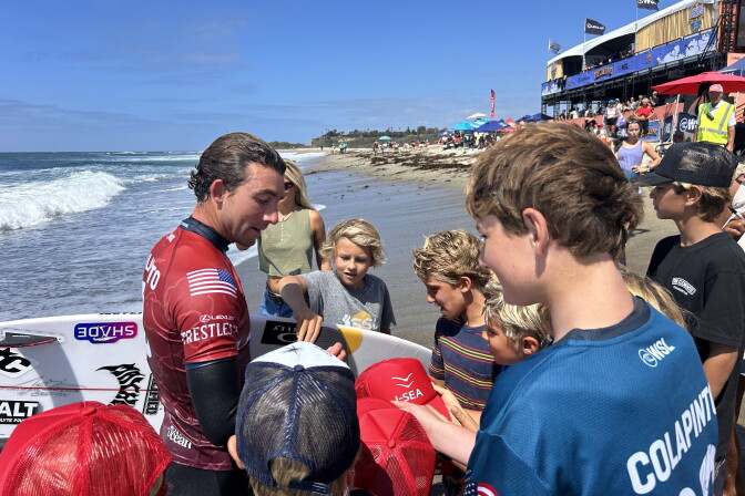 A wetsuit-clad surfer signs hats and a surfboard for a group of children gathered on the beach near a competition venue. One of the kids has a shirt that reads "Colapinto" on the back. 