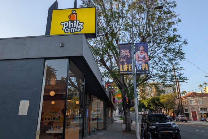 The photo shows the exterior of a Philz Coffee shop, easily identifiable by a large yellow sign with the Philz Coffee logo. The shop features large windows, allowing a view inside where you can see hanging lights and tables.