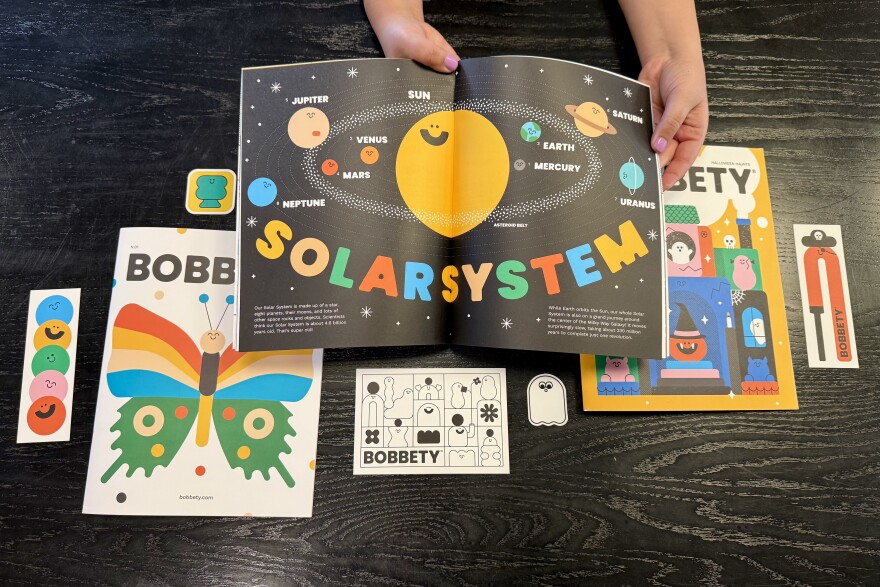 A pair of hands with medium light skin tone hold open a brightly colored magazine depicting the solar system with a bright yellow circle surrounded by smaller circles in different colors. The magazines in the background have a butterfly and ghosts on the cover. 