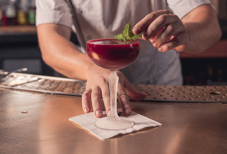 a hand pushes forward a champagne coupe filled to the brim with a dark red drink