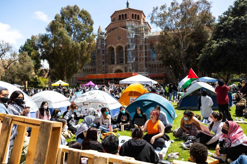Various people sit in a circle on a grassy lawn in front of a large brick university building. Many wear surgical masks and keffiyehs. There are tents set up around them and a Palestinian flag can be seen hanging from the top of one of the tents.