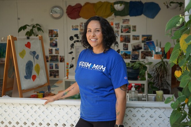 A woman with a medium skin tone and a bright blue t-shirt stands in front of an open-air classroom smiling. She has short, curly dark brown hair. Behind her is an easel with a child's painting. Further back there are photos and a clock on a wall. To her right, a tangle of green leaves poke into the frame. 