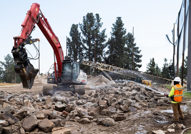 A person in a hard hat and orange vest sprays water onto a pile of rubble while a big machine works nearby. 