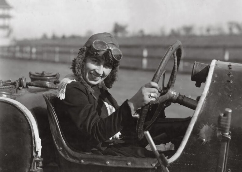 A black and white photo of a fair-skinned woman with driving goggles on the top of her head, behind the wheel of a car