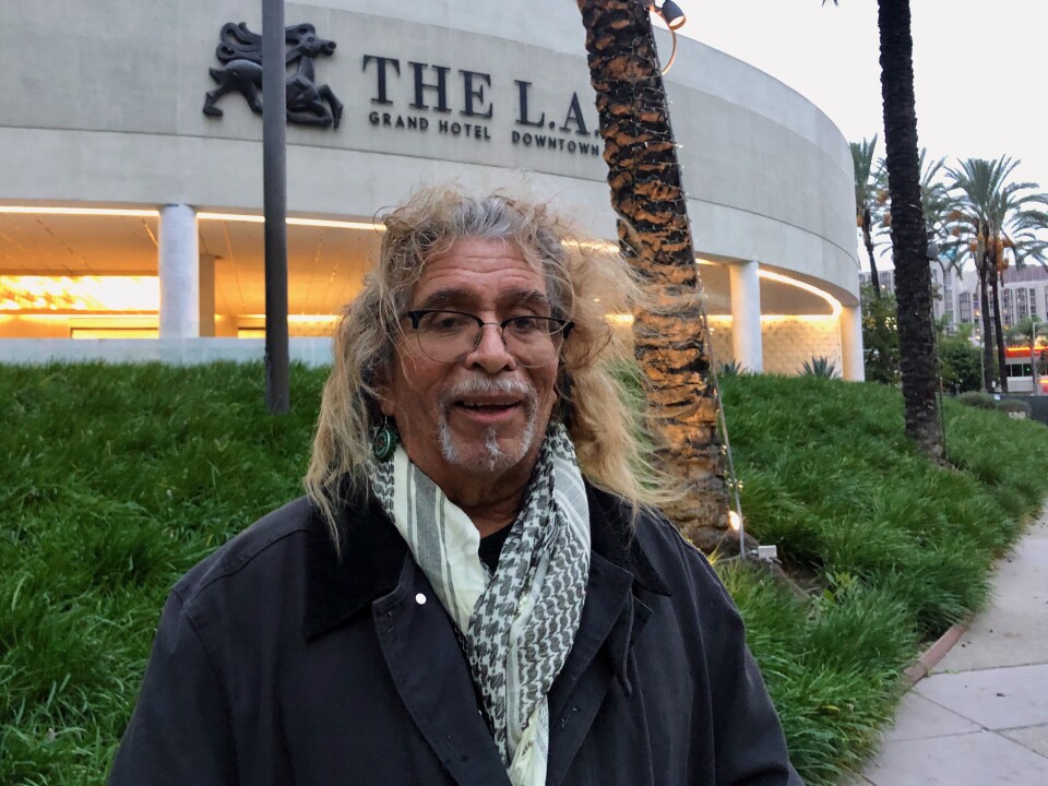 A man with medium-ton skin with long hair and a gray goatee stands in front of the L.A. Grand Hotel in downtown Los Angeles