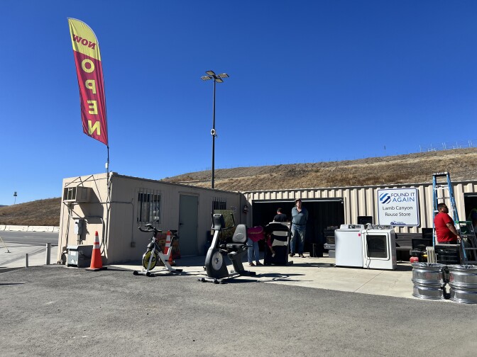 Three people are walking around an outdoor space on a bright, sunny morning. There's a variety of objects displayed and scattered on the pavement and asphalt in front a tan, metal structure. The objects include a pair of exercise bikes, a washer and dryer, and a large teal ladder propped up against the wall.