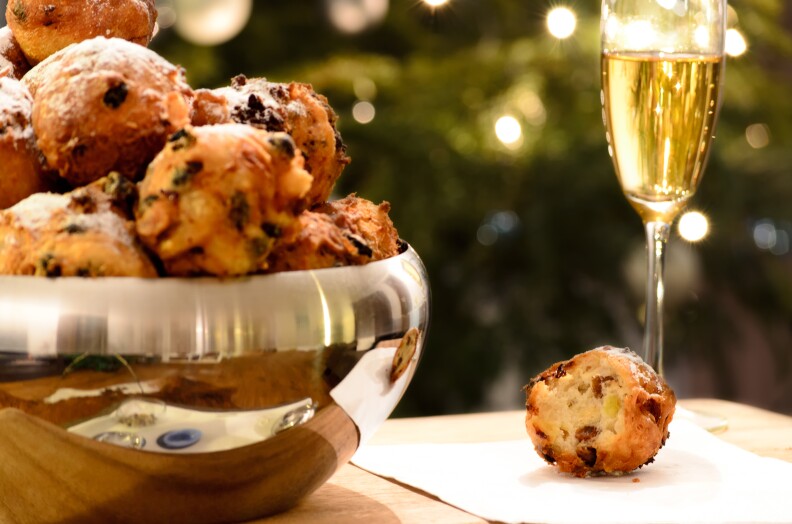 A photograph of a festive holiday scene: There is a pretty, metal bowl laden with balls of golden-brown, deep fried fritters with raisins. The fritters have been topped off with a dusting of confectionary sugar. To the right of the bowl, one of the fritters sits on a napkin, a bite has already been taken out of it. And next to that morsel, there is a glass of champagne, sparkling in the glow of the holiday lights in the background.