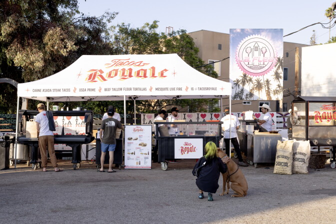 A white pop-up tent reading "Tacos Royale" in red-and-yellow lettering set up in a parking lot, with staff cooking behind counters, bags of mesquite charcoal, and a customer crouching beside a dog.