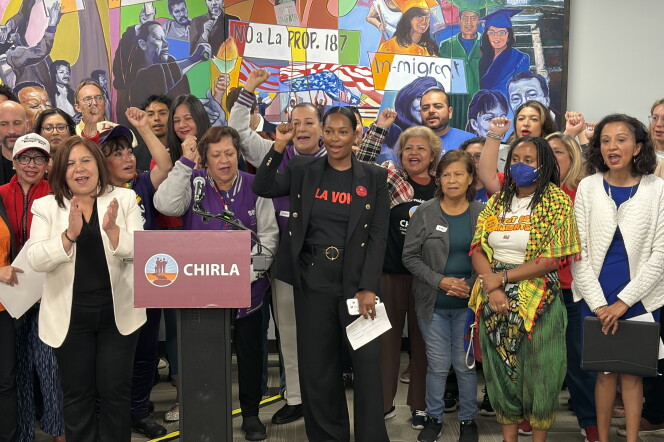 A group of people stand behind a podium with a sign that reads "CHIRLA." They also stand in front of a painted mural. Some people are raising a fist upend others are clapping. 
