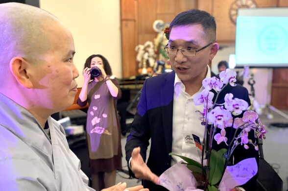 An Asian American man in his 40s wearing glasses and a suit (l.) presents a potted plan to a nun (r.) who wears a smile and a shaven head.
