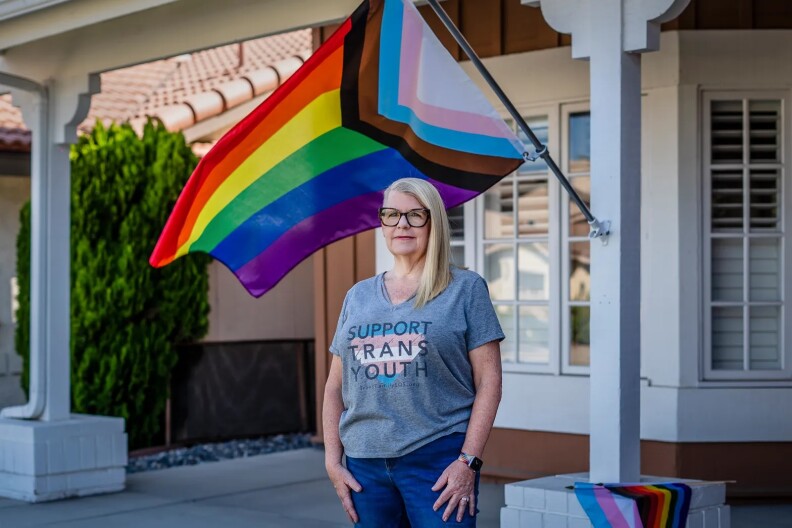 A female-presenting person with light skin tone and blonde hair is wearing a shirt saying 'Support Trans Youth' and standing in front of a pride flag