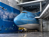 The front 70-feet of a Boeing 747 aircraft appears to be sticking out of a wall in a large room. The plane is painted mostly blue with a white underbelly and the "Korean Air" logo on the left side. A wall painted to look like the blue sky with white clouds and various small aircraft images is to the right of the plane