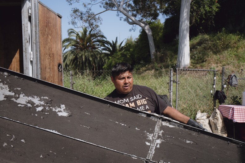 A medium brown Latino man with dark hair and a black shirt unloads something from a truck.