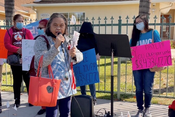 A woman of Asian descent with short gray hair speaks into a microphone on a sidewalk outside the fenced yard of a stucco house. Several other women stand in the background with signs. One pink sign reads "Pay up!"