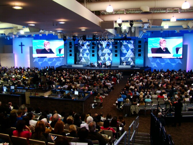 A large theatre style room has people filling most rows. At the front large screens project a woman on stage with a panel. A cross is visible at the left.