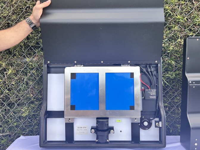 A man's hand, with a dark metal watch on the wrist, is holding up the metal front of a battery system to show off the parts inside. The internal pieces are made out of metal, screws, and minimal wiring. 