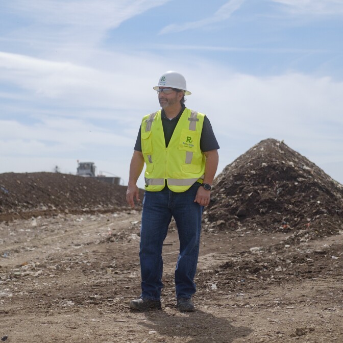 A man with a white hard hat and neon safety vest stands in the foreground. Piles of dirt stretch out behind him. 