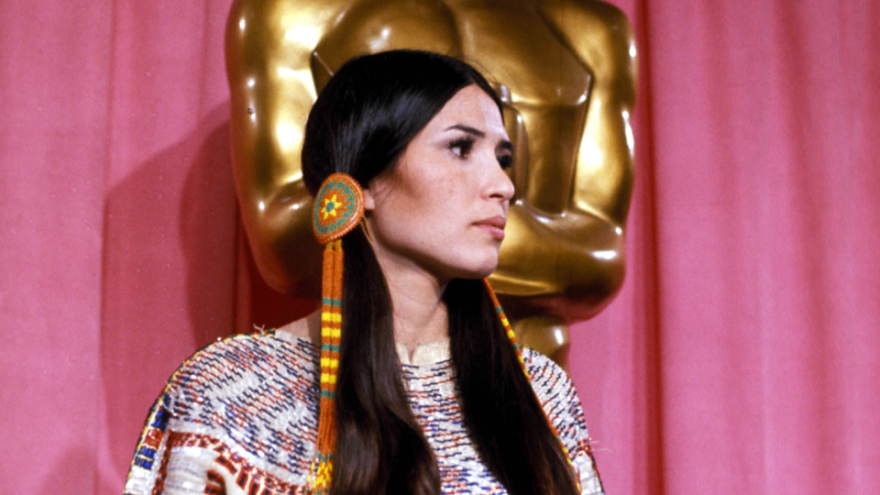 An image of Native American woman Sacheen Littlefeather, wearing tradition clothing. She stands in front of a large golden Motion Picture Academy Oscar statue.