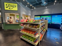 The interior of Tiny's showing shelves of imported snacks including Japanese Kit-Kats and Korean chips, with the order counter and illuminated Tiny's sign visible in the background.