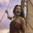 A female-presenting person wearing a terracotta strapless top and a necklace stands holding the rope of a sail. The sail and a cloudy sky are visible in the background.