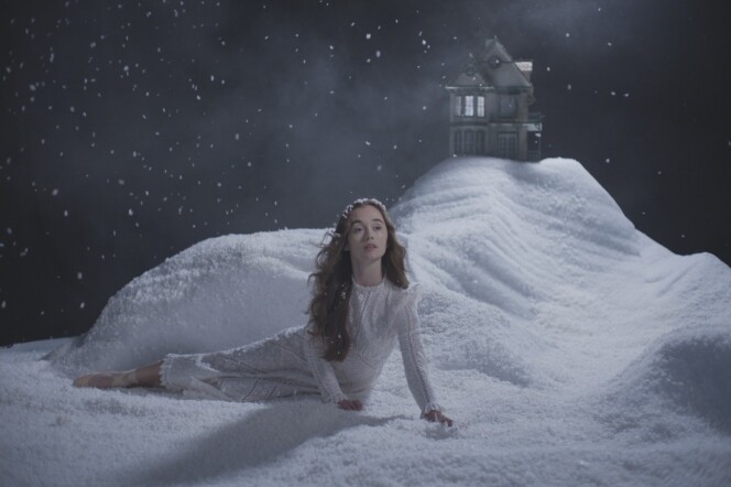 A woman in a white gown lies on a white snow-covered stage scene with a house in the background. 